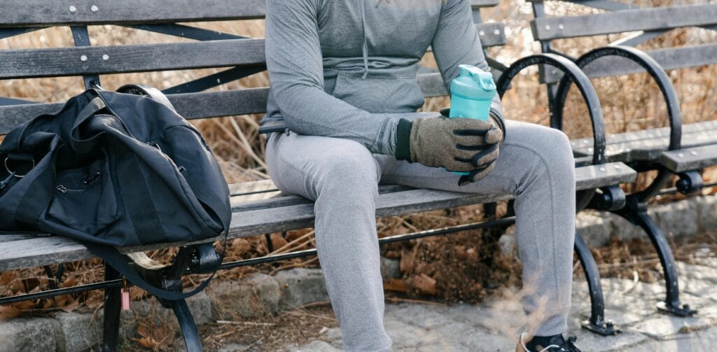 A man in activewear rests on a park bench holding a shake bottle, enjoying a break.
