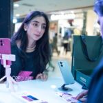 Woman at smartphone demo counter engages with salesperson in Islamabad retail setting.