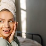 A woman with a towel wrapped around her head applying under-eye masks in a sunlit room.